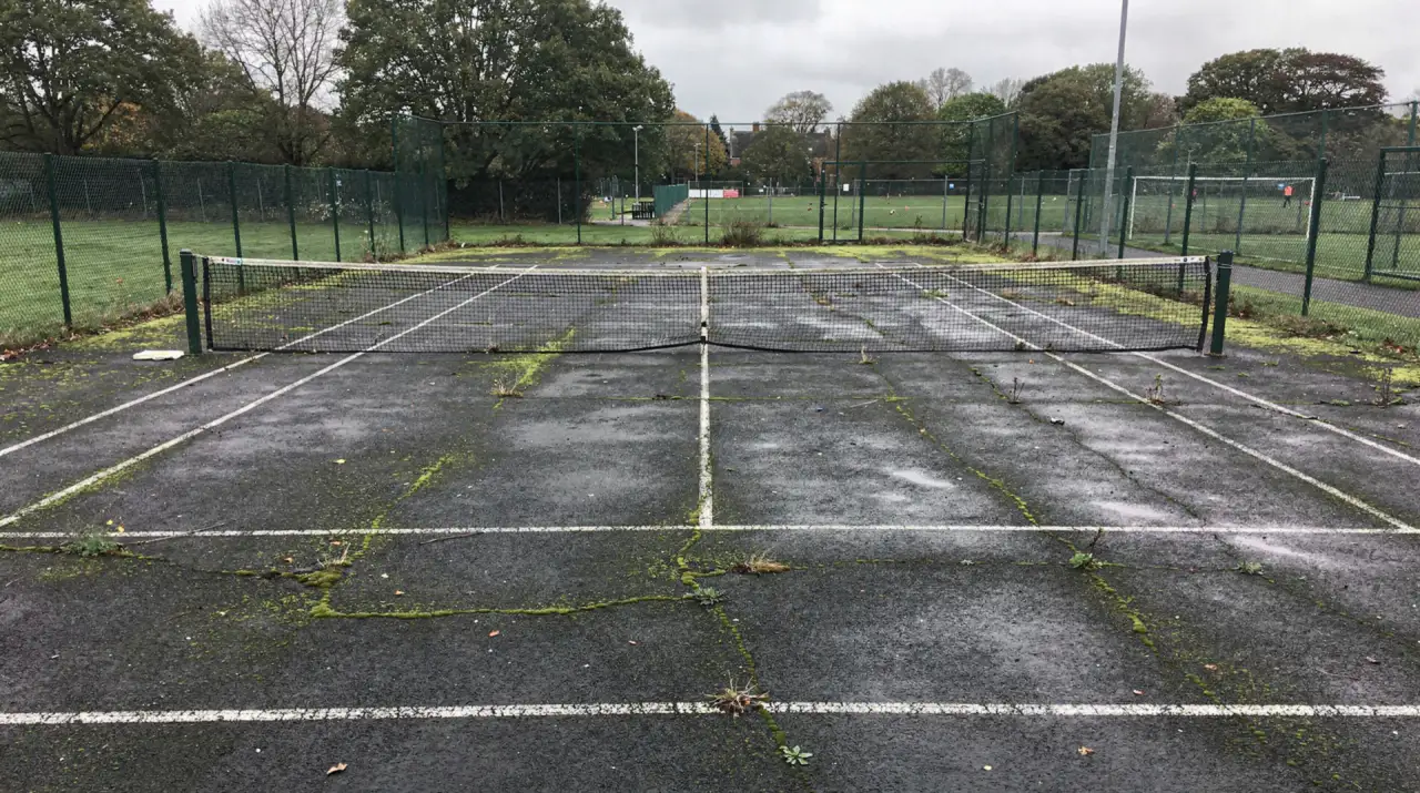 A British school tennis court in plain tarmac, its white lines faded and moss spreading through the surface cracks, of the kind restored by specialist court cleaning
