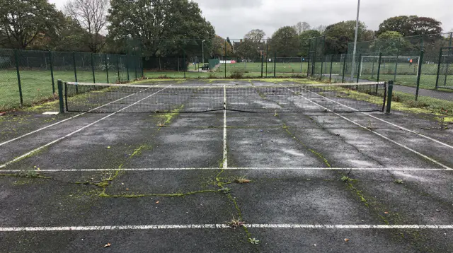 A British school tennis court in plain tarmac, its white lines faded and moss spreading through the surface cracks, of the kind restored by specialist court cleaning